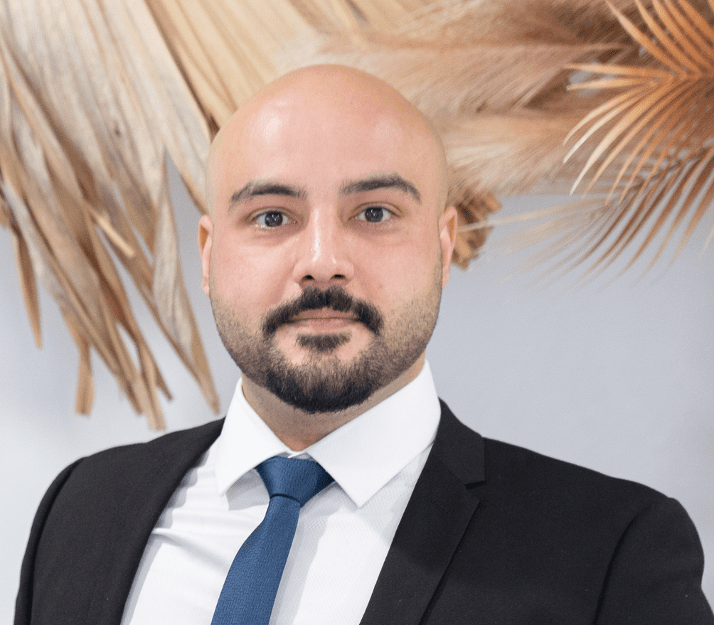 Ridha Hussain Profile Image A man in suit and tie standing next to palm trees.